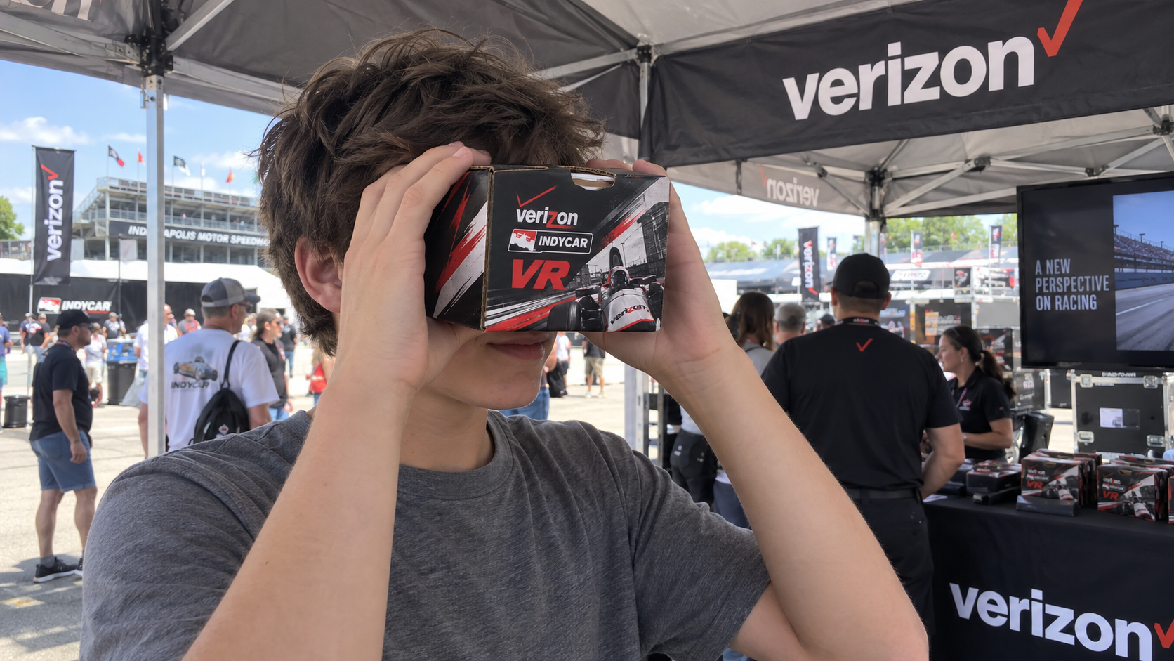A fan holds an IndyCar-branded cardboard VR viewer to their eyes in front of the Verizon tent